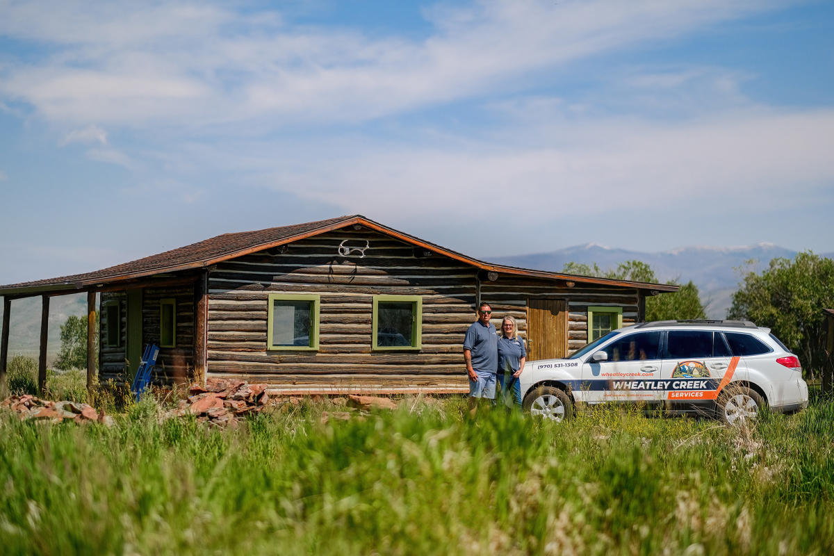 Wheatley Creek Services owners standing in front of a historic Granby Colorado log cabin with their branded company vehicle